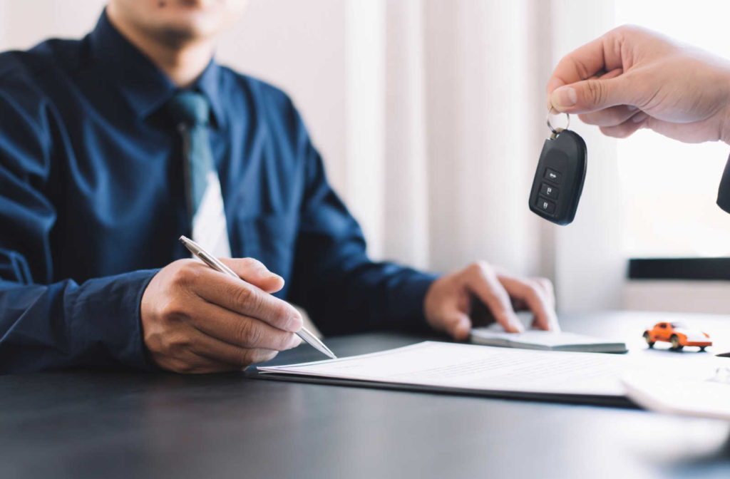 A male signs a piece of paper to purchase a vehicle using a vehicle loan while a hand holds the keys out in front of him.