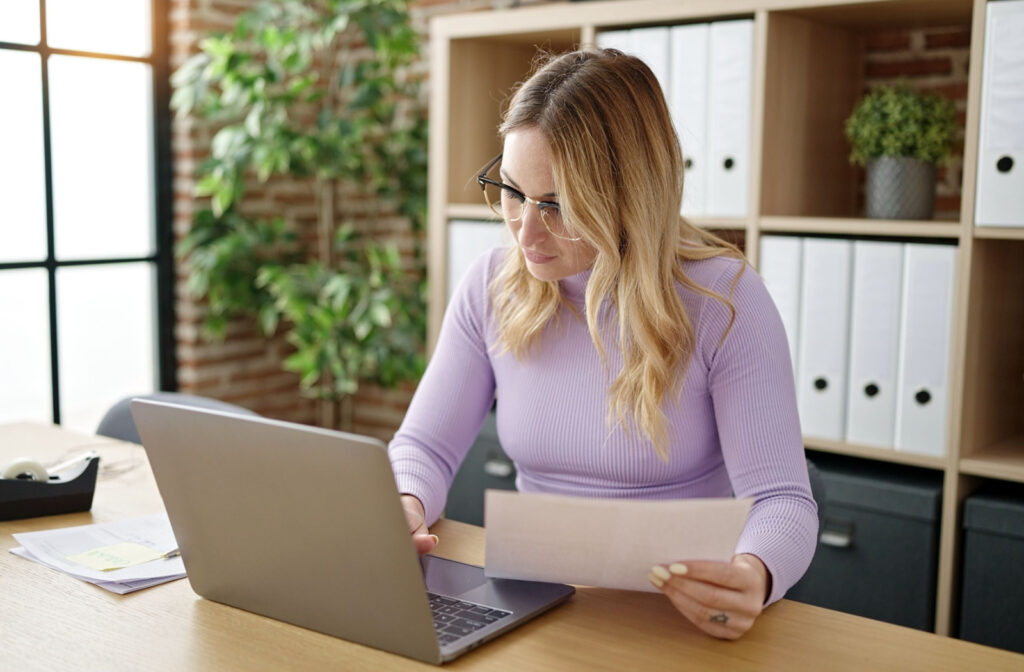 A young woman reviewing her payday loan contract while holding the paperwork in her left hand and using the laptop with her right hand.