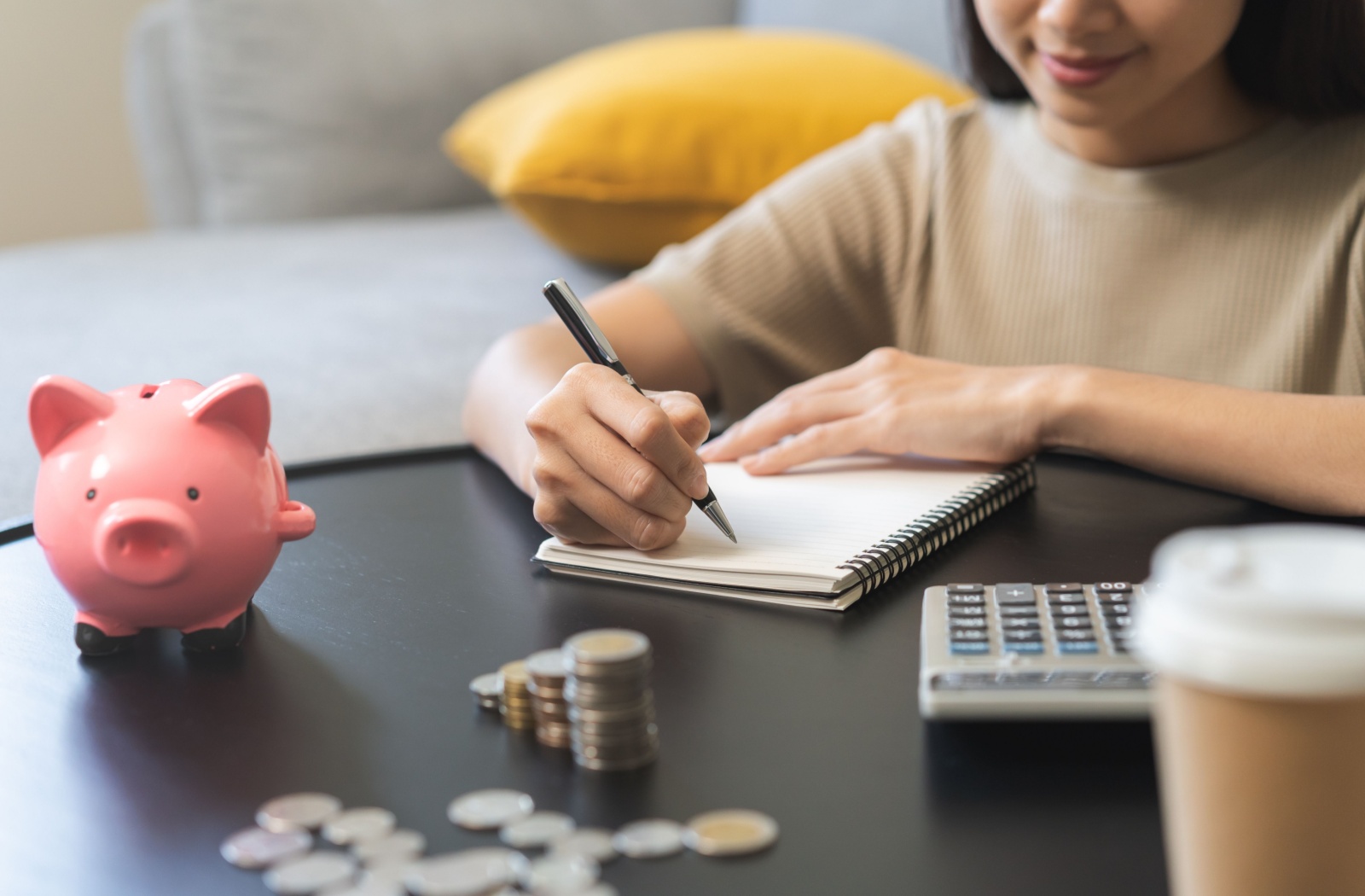 Close-up view of a desk with a piggy bank, coins, a calculator, and a notebook on it as a person in the background writes in the notebook.