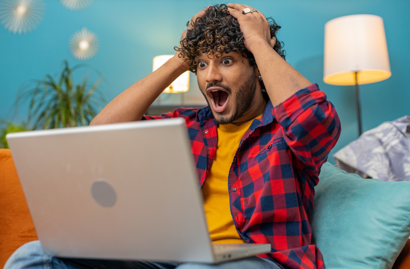 A person sitting on their couch, looking at their laptop in shock and excitement with their hands on their head because they won the lottery.