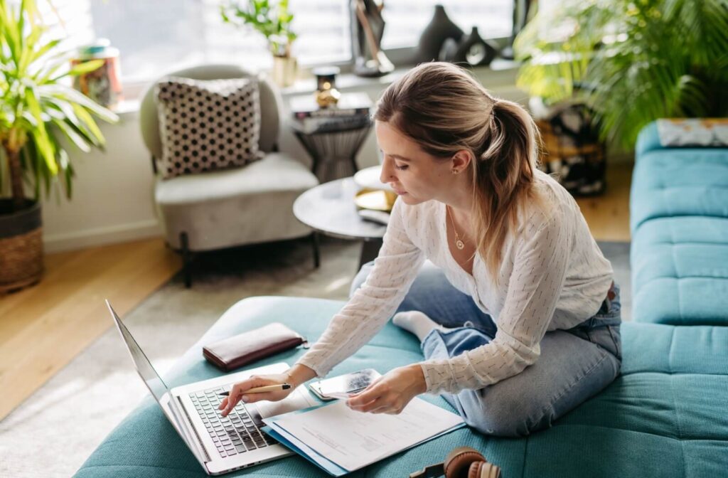 A person sitting on a turquoise couch at home and using their laptop and a notepad to tally up and pay off their debt.