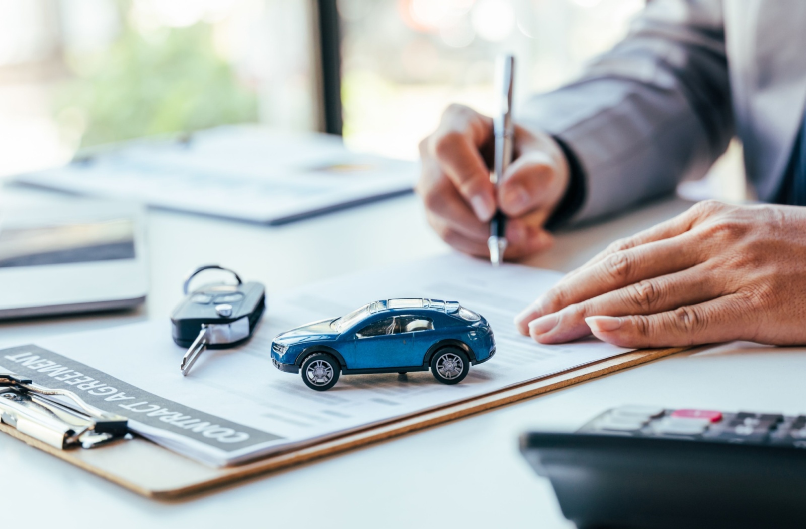 Close-up of a person sitting at a desk signing a new loan agreement to refinance their car. A set of car keys and a blue toy car sit on the table.