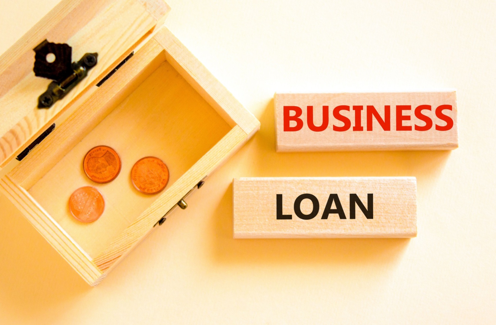 Wooden blocks labeled “BUSINESS” and “LOAN” beside an open box containing coins, symbolizing small business financing or funding options.