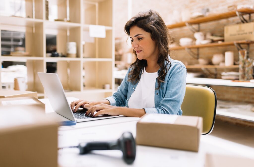 A small business owner managing online orders from a workspace, typing on a laptop surrounded by shipping boxes and a barcode scanner.