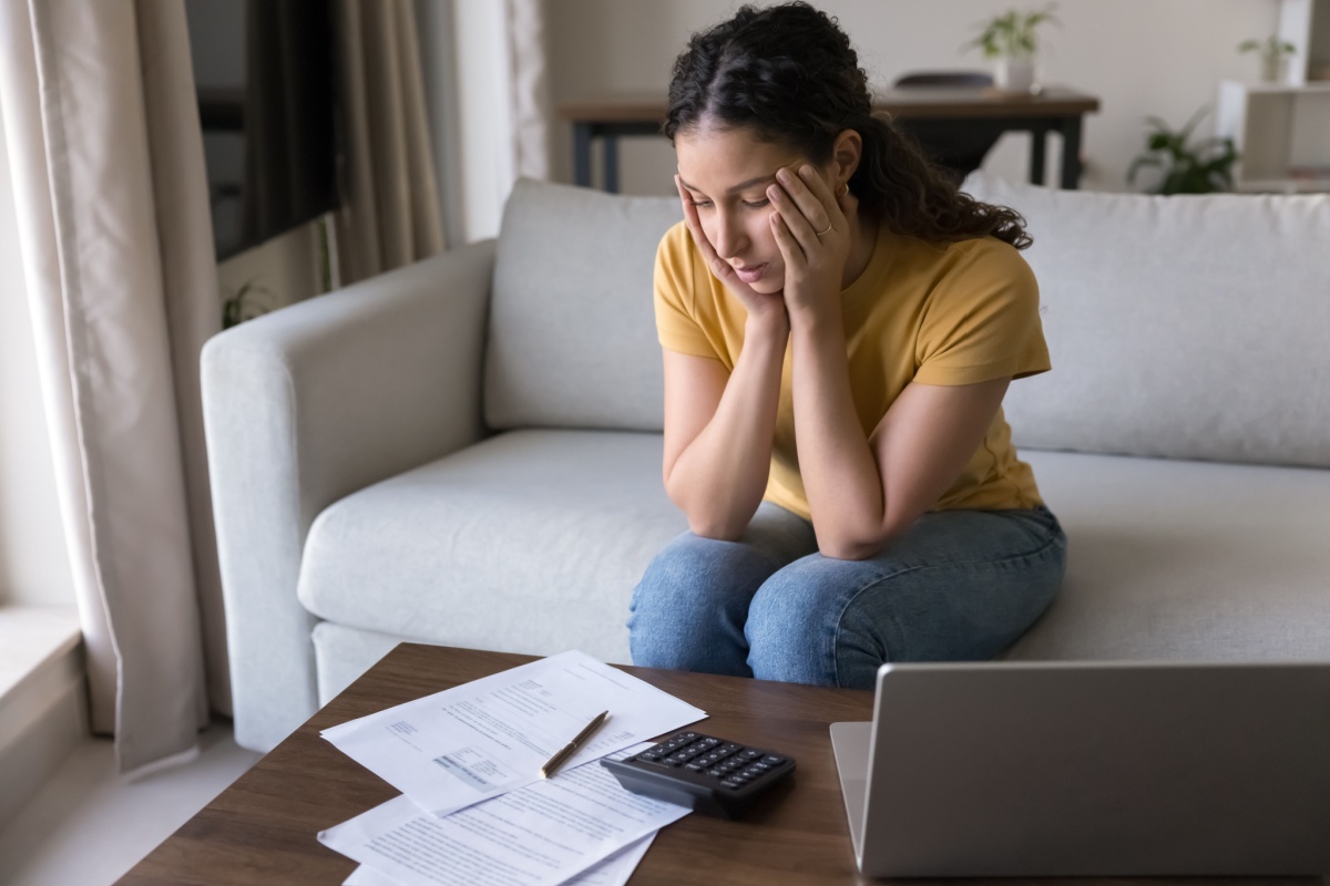 A person sitting on a coach with their hands on their cheeks, looking down at loan paperwork, stressed.