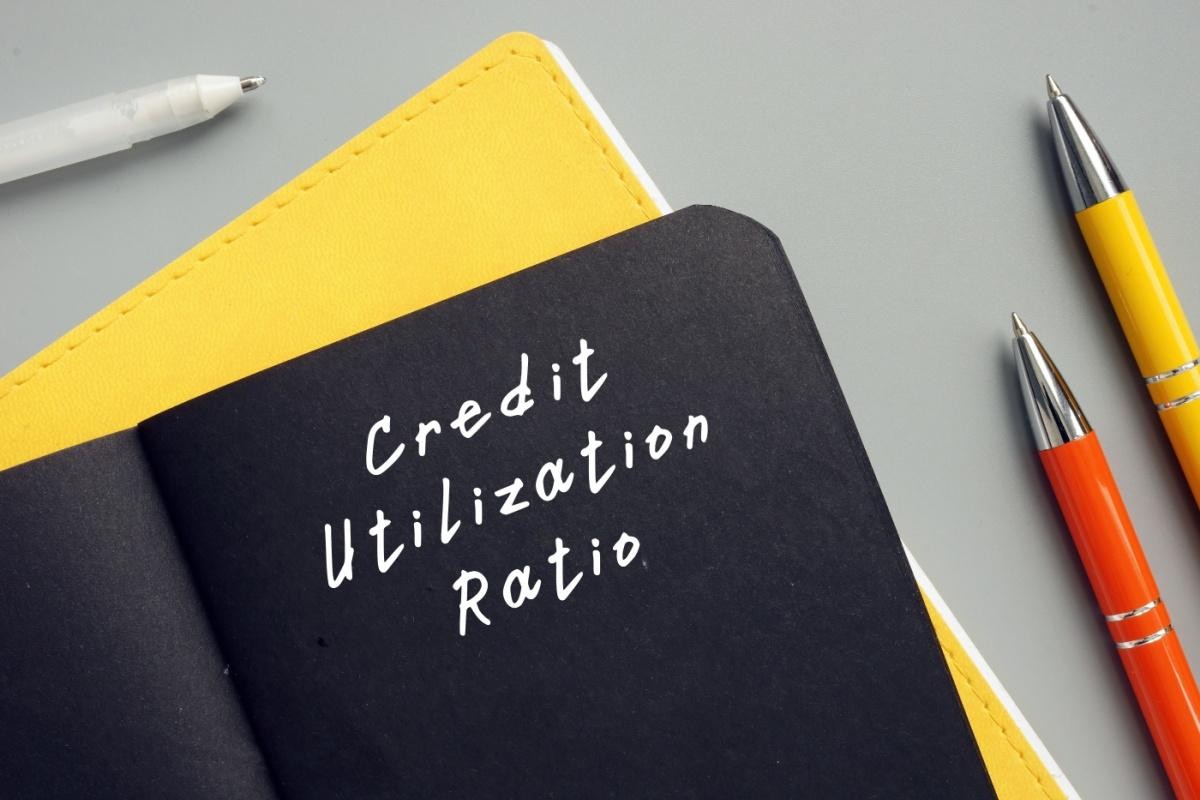 A top-down view showing a black notebook with the words 'Credit Utilization Ratio' written in white text, resting on a yellow notebook on a gray surface with three pens of different colors nearby, symbolizing financial planning and debt management.